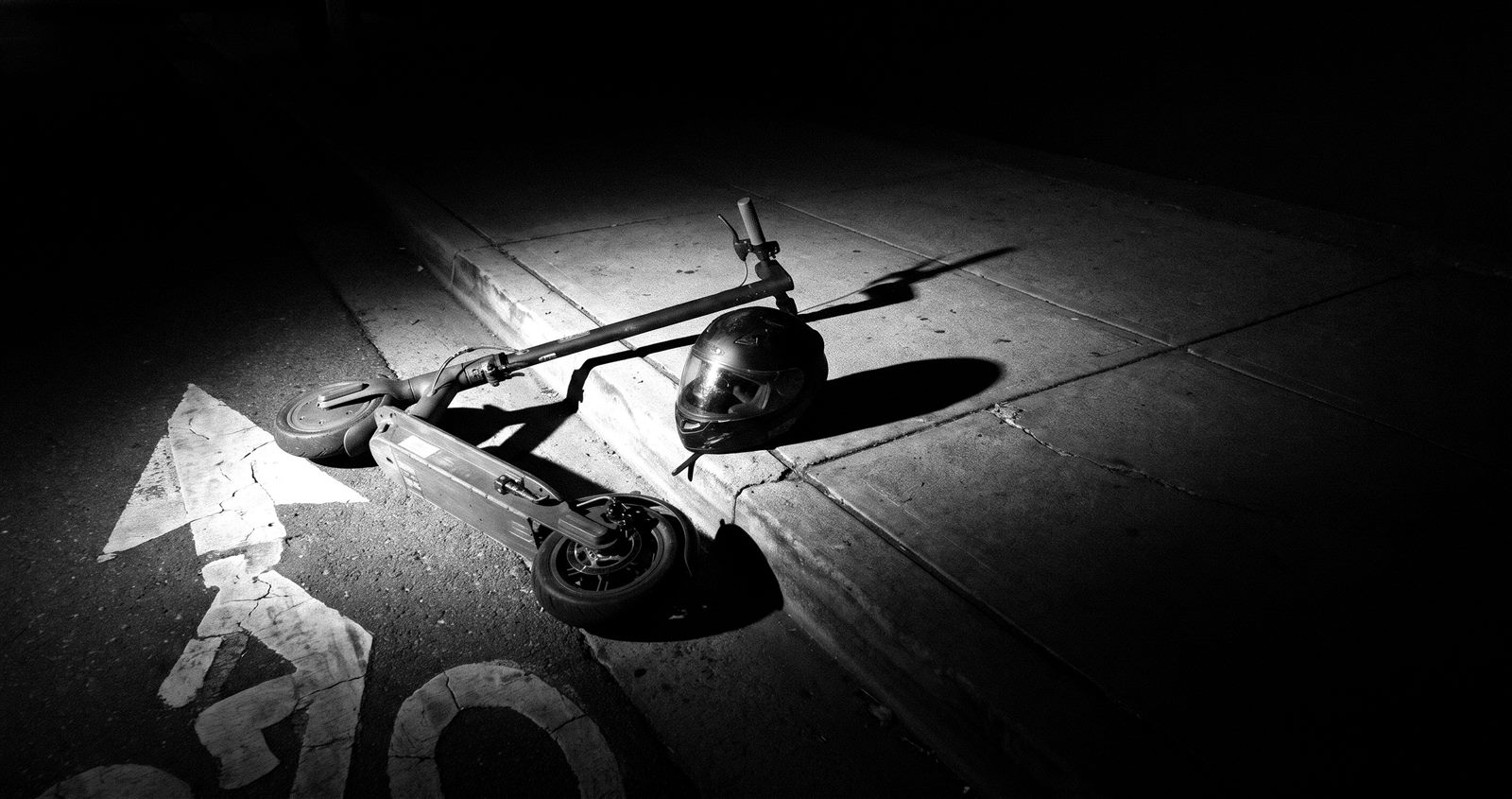 An electric scooter and a helmet lying at the edge of a sidewalk at night